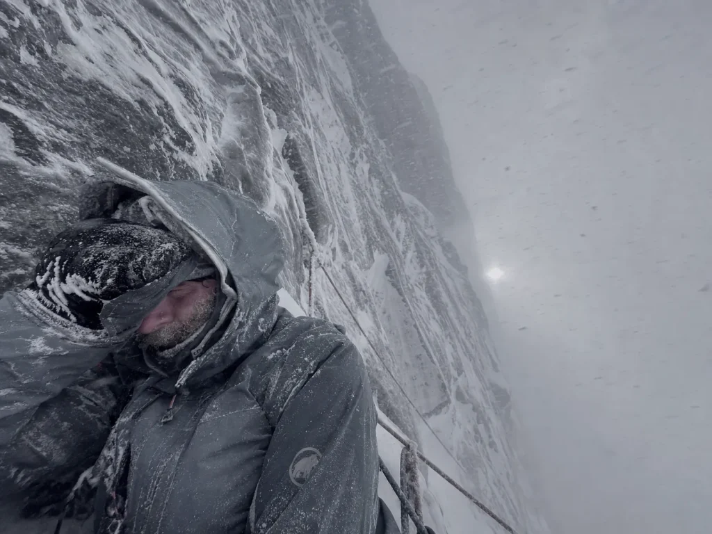 Climber hunkered against wind and spindrift on an exposed Eiger stance in a storm