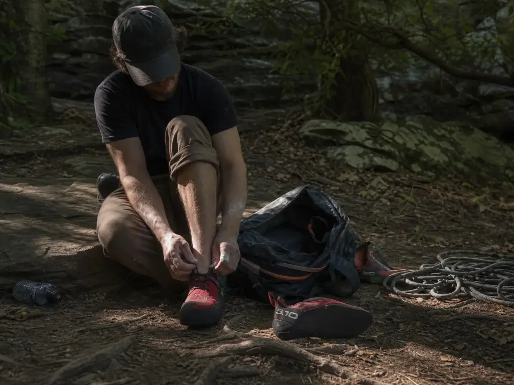 Climber lacing a trad shoe at a crag base with a slipper and a Velcro shoe beside the pack